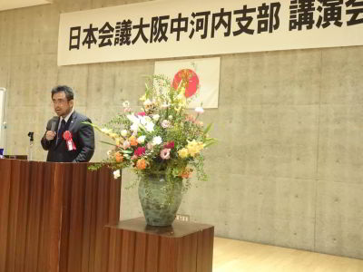 スーツを着た男性が壇上でマイクを持って話している。