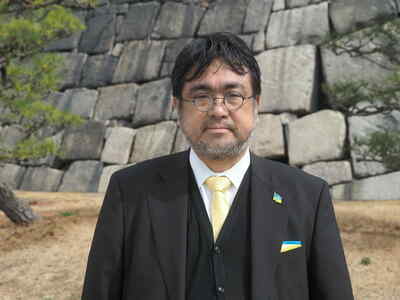 黒いスーツに白いネクタイをしている。髭を生やし、メガネをかけている40代の男性が正面を向いて微笑んでいる。